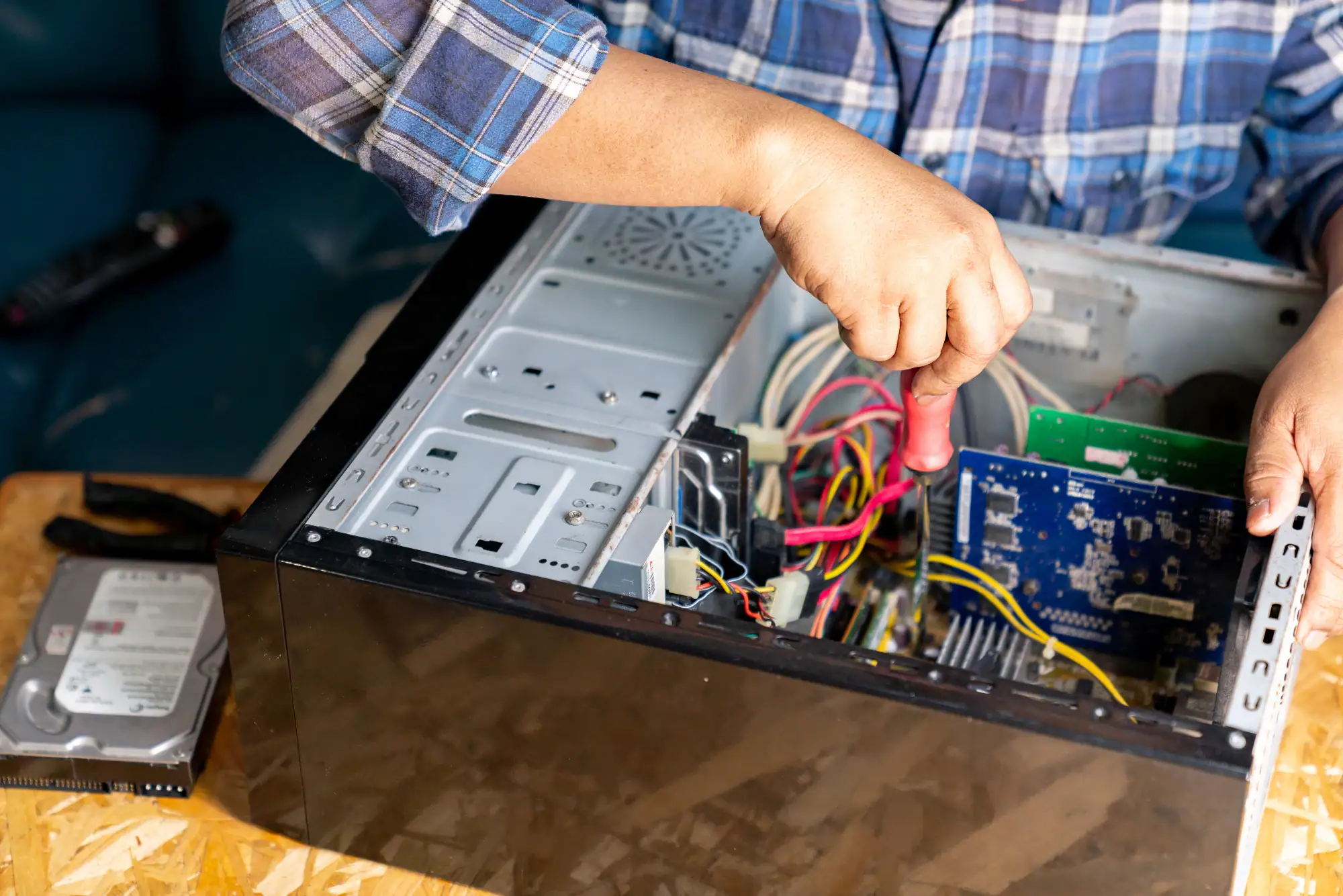 A person in a plaid shirt uses a screwdriver to work inside an open desktop computer case; various cables and computer components are visible, and a hard drive lies on the table nearby—typical tasks for Electrical Services Cook and Will County, IL.