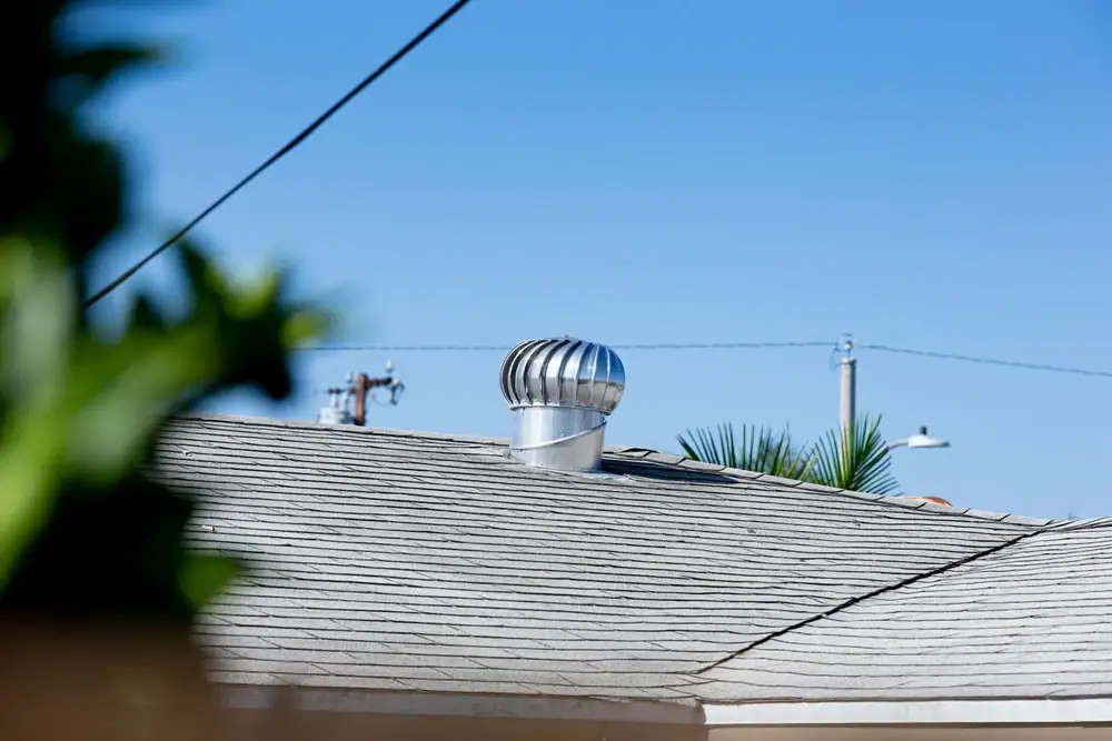 A silver turbine vent is installed on a shingle roof in IL, with a clear blue sky and utility poles—reminders of trusted electrical services in Cook and Will County—visible in the background. Some green leaves are blurred in the foreground.