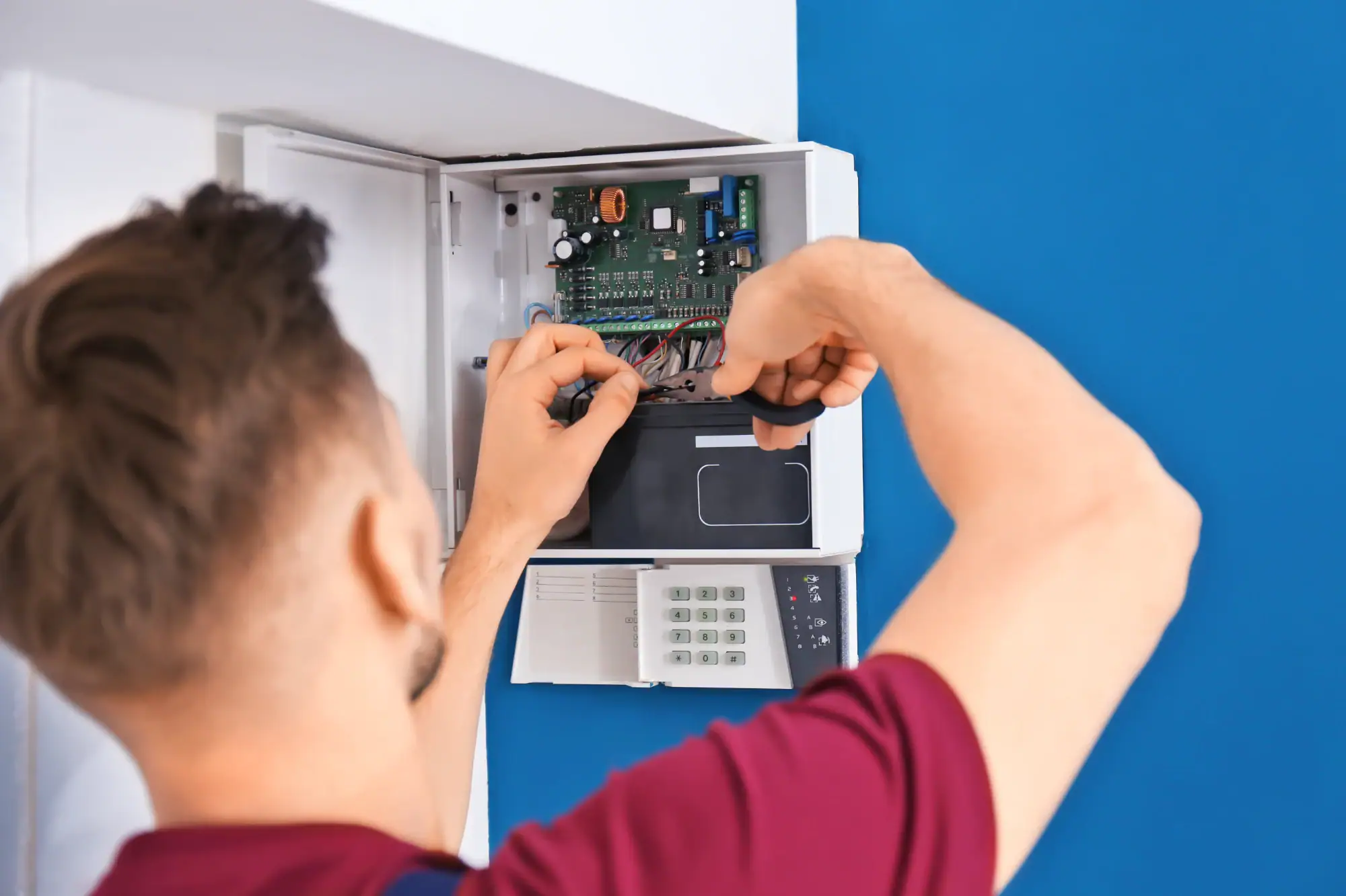 An electrician Cook and Will County in a red shirt is working on the wiring inside an open security alarm control panel mounted on a blue and white wall.