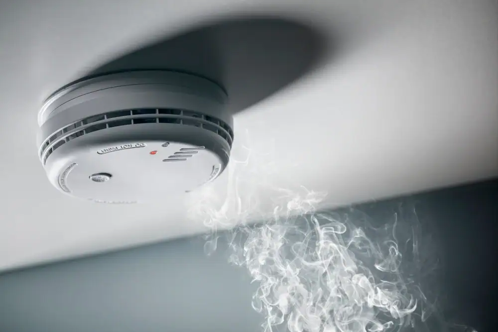 A smoke detector mounted on a ceiling with smoke rising toward it, indicating the presence of smoke or a potential fire—trust an electrician Cook and Will County, IL, for installation and safety checks.