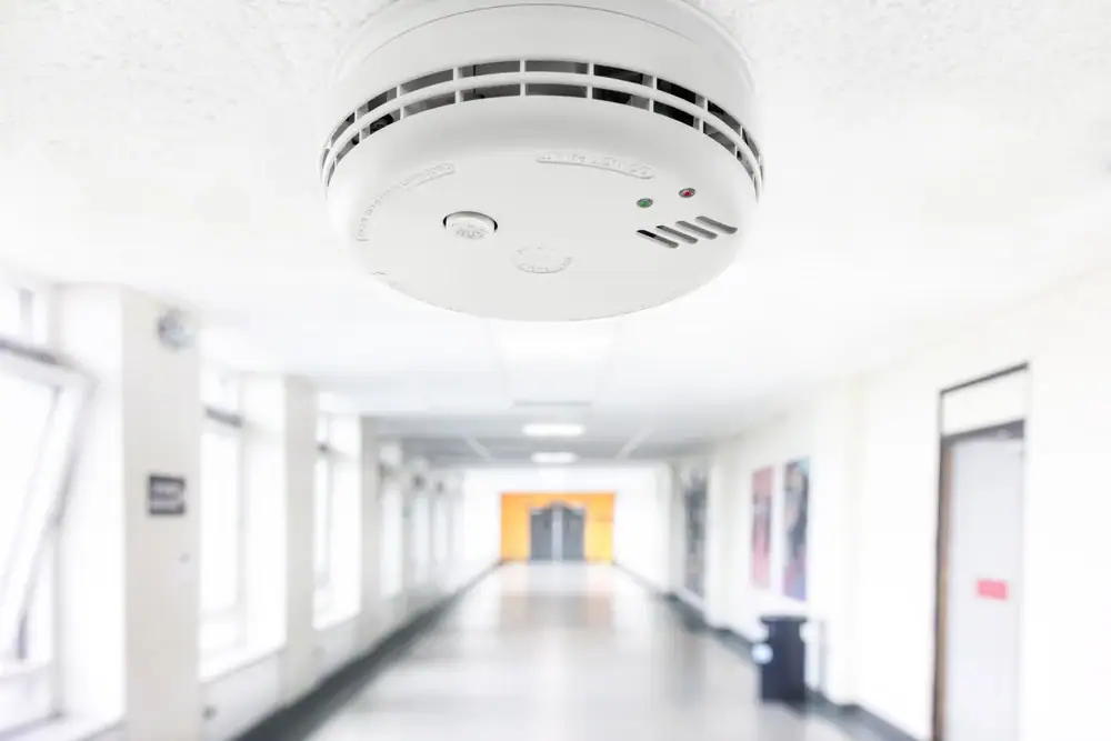 A close-up of a smoke detector mounted on a white ceiling in a brightly lit corridor, showcasing the quality work of an electrician Cook and Will County, IL, with windows and doors visible along the hallway.