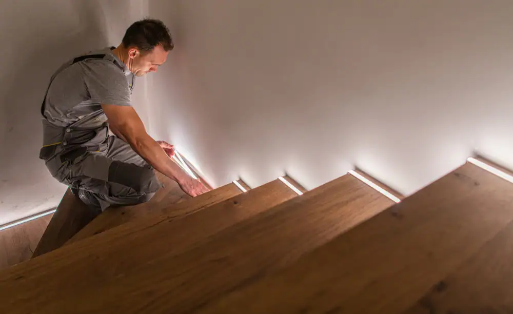 An electrician in Cook and Will County installs LED strip lights along the side of wooden stairs, adding modern illumination to the staircase in a stylish interior setting.