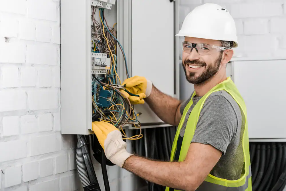 A smiling electrician wearing a white helmet, safety glasses, yellow vest, and gloves works on an open electrical panel with exposed wires in a white brick room in Chicago, IL