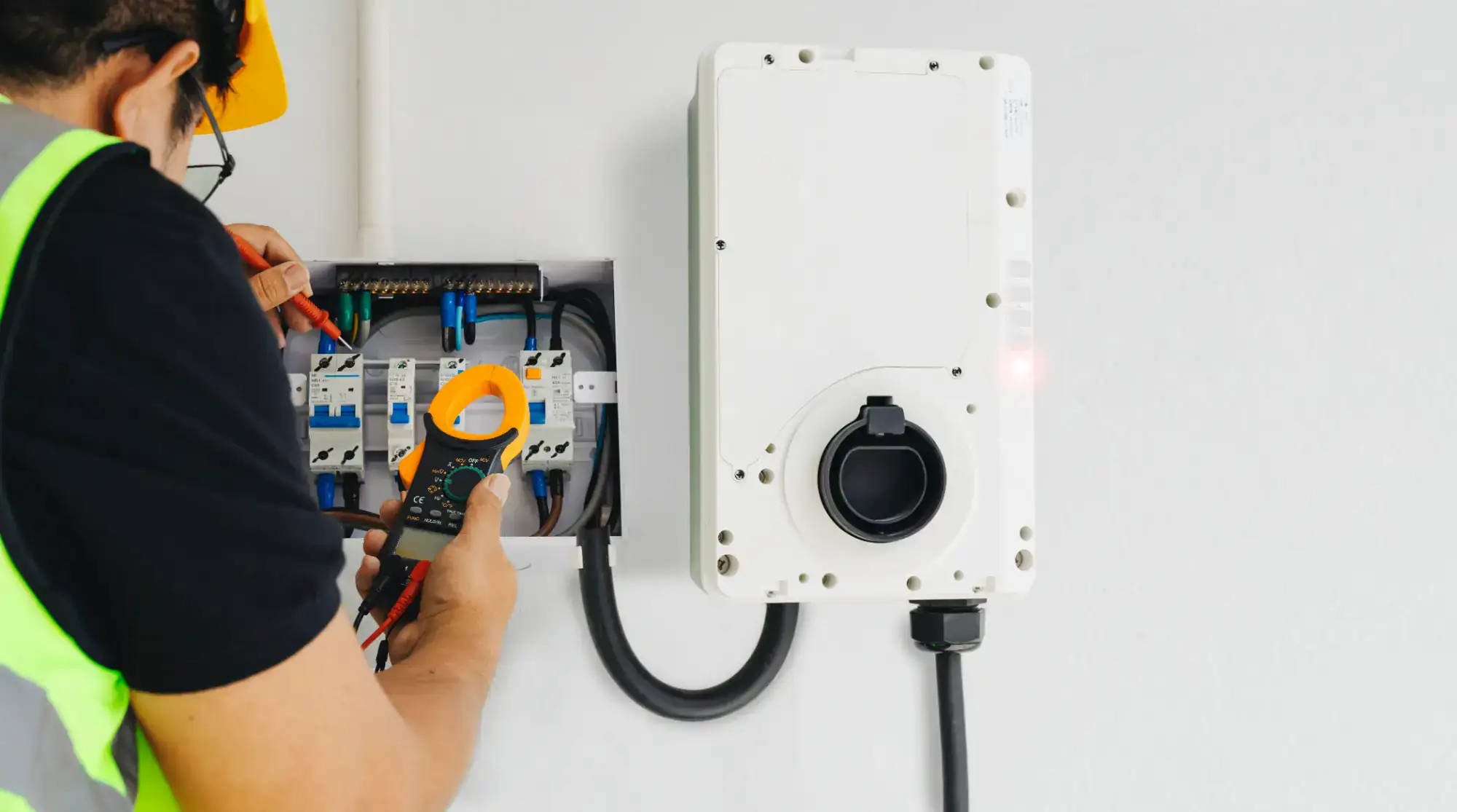 An electrician in a yellow hard hat and safety vest tests electrical wires inside a circuit breaker panel on a wall, using a digital clamp meter; a white control box is mounted nearby.